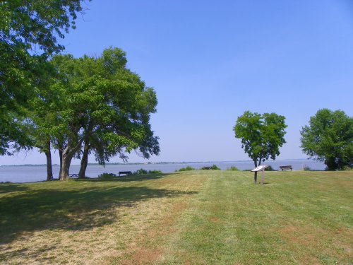 A serene landscape featuring green grass, trees, and a calm river under a clear blue sky.