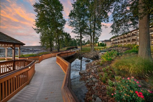 A scenic walkway by a calm waterway, surrounded by trees and colorful flowers, with a building in the background at sunset.