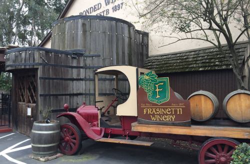 A vintage truck with a winery sign parked outside a rustic building, surrounded by barrels and greenery.