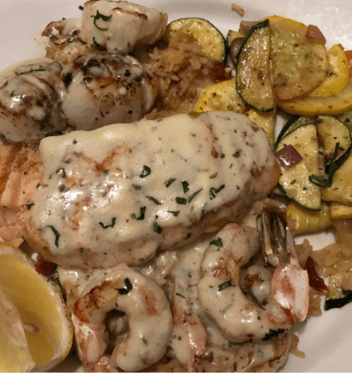 A plate of seafood featuring grilled fish, shrimp, scallops, rice, and sautéed zucchini and squash, garnished with lemon.