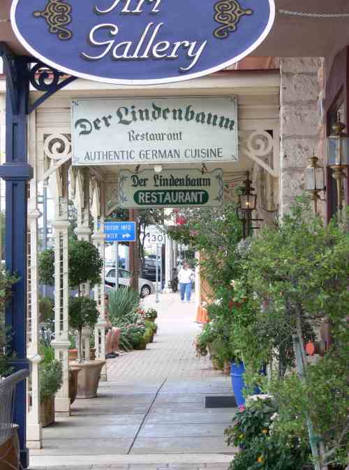 A charming street view featuring an art gallery and a restaurant sign for "Der Lindenbaum" with greenery lining the walkway.