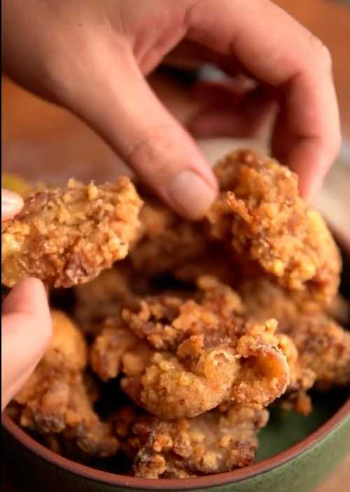 A hand holding crispy fried chicken pieces from a bowl, showcasing a golden-brown, crunchy texture.