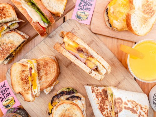 A wooden board with various grilled sandwiches and a drink, featuring melted cheese and colorful toppings.