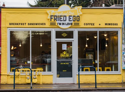 Bright yellow storefront of "Fried Egg" café, featuring breakfast sandwiches, coffee, and mimosas. Large windows and inviting entrance.