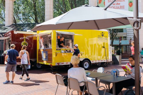 A vibrant yellow food truck serves customers in a sunny plaza, with people dining at nearby tables.