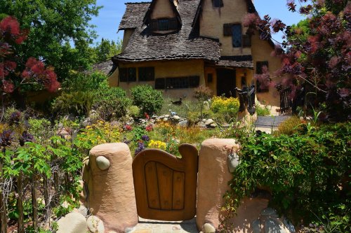 A whimsical cottage with a thatched roof surrounded by colorful flowers and lush greenery. A charming wooden gate is visible.