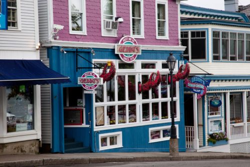 A colorful storefront with pink and blue accents, featuring flower baskets and large windows, in a quaint town setting.