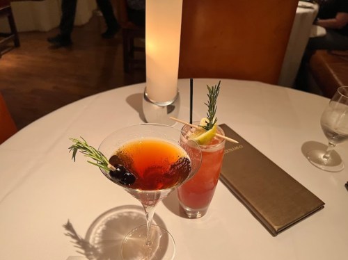 Two cocktails on a white tablecloth: a martini with rosemary and blueberries, and a drink with a lime wedge and rosemary.