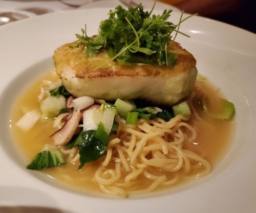 A plate of noodles topped with a piece of fish and garnished with fresh herbs and vegetables in a light broth.