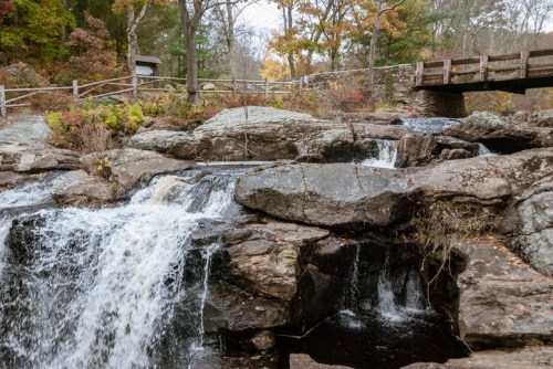 A serene landscape featuring a waterfall cascading over rocks, with autumn foliage and a wooden bridge in the background.