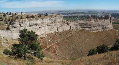 A panoramic view of a rocky cliff with sparse vegetation, overlooking a valley and distant cityscape under a clear blue sky.