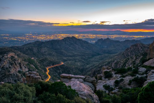 A scenic view of mountains at sunset, with city lights twinkling in the valley below and a winding road visible.
