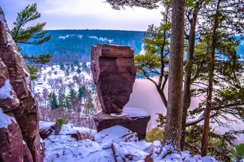 A snow-covered rock formation stands on a cliff overlooking a serene lake surrounded by trees.