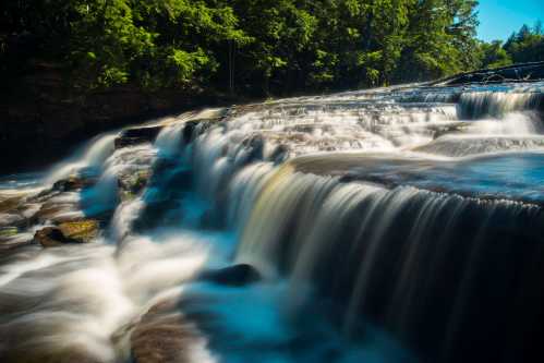 A serene waterfall cascades over smooth rocks, surrounded by lush green trees under a clear blue sky.