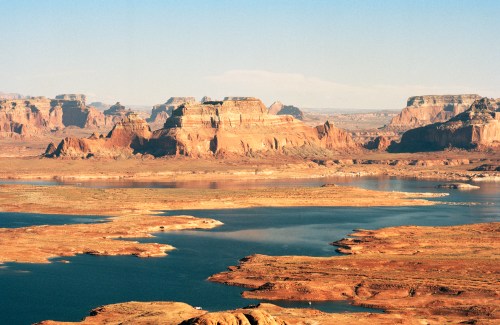 A vast desert landscape with red rock formations and a blue lake, under a clear sky.