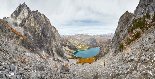 A panoramic view of rugged mountains and a serene lake, with a lone hiker in the foreground amidst rocky terrain.