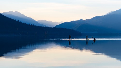 Two paddleboarders glide across a calm lake, surrounded by misty mountains and a serene, reflective surface.
