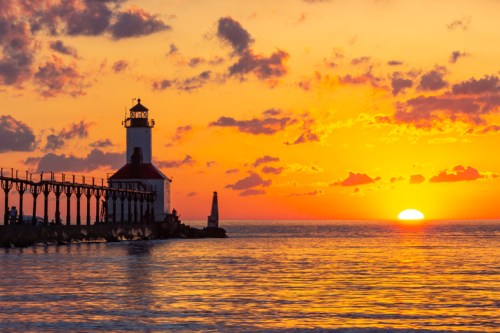 A lighthouse stands by the water at sunset, with vibrant orange and purple skies reflecting on the calm sea.