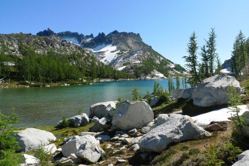 A serene mountain landscape featuring a clear lake, rocky shore, and evergreen trees under a bright blue sky.