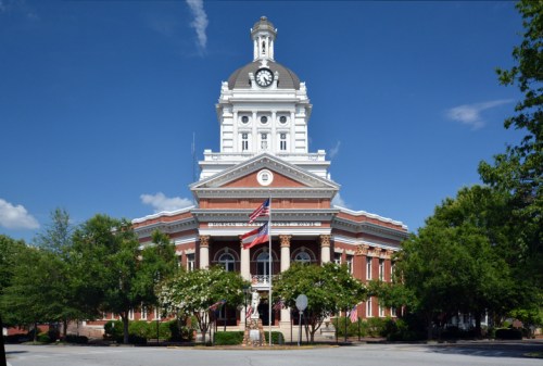 Historic courthouse with a clock tower, surrounded by trees and a clear blue sky, featuring an American flag in front.