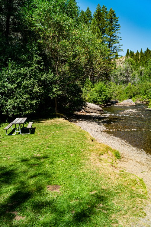A serene riverside scene with a picnic table, lush greenery, and tall trees under a clear blue sky.