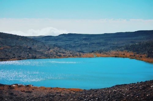 A serene blue lake surrounded by rocky terrain and distant hills under a clear sky.