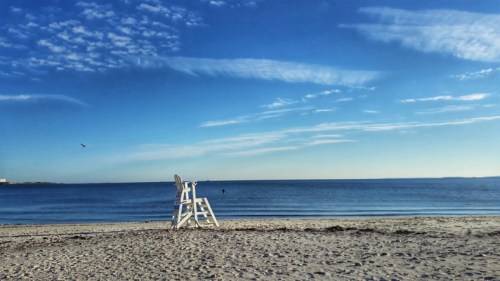 A white lifeguard chair on a sandy beach with calm blue water and a clear sky in the background.