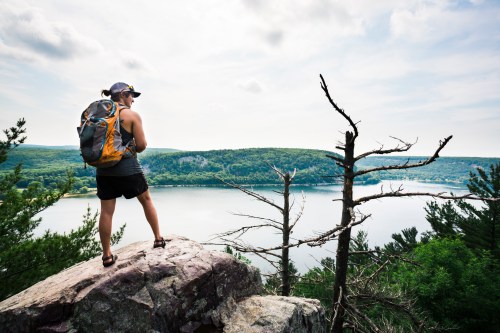 A hiker stands on a rock overlooking a lake surrounded by lush greenery and distant hills under a cloudy sky.