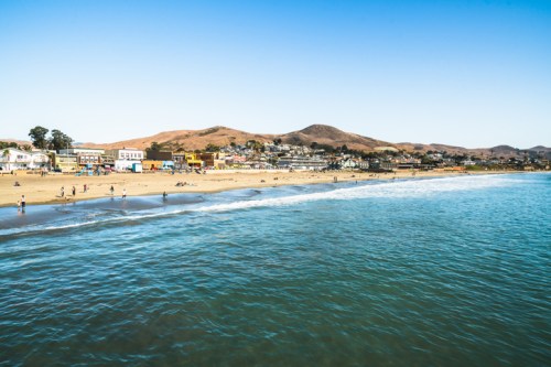A scenic beach view with gentle waves, sandy shore, and distant hills under a clear blue sky.