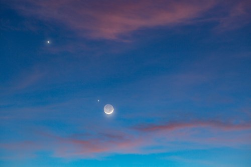 A crescent moon and two bright planets in a colorful twilight sky with soft clouds.
