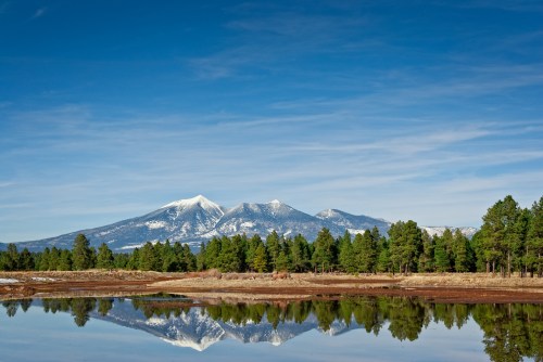 Snow-capped mountains reflect in a calm lake, surrounded by green pine trees under a clear blue sky.