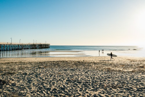 A serene beach scene with a pier, surfers in the distance, and a clear blue sky reflecting on calm waters.