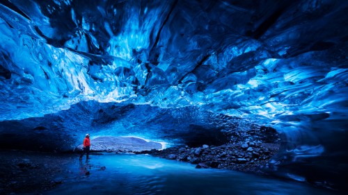 A person stands in an icy cave with stunning blue ice formations overhead and a river flowing nearby.