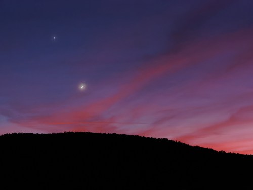 A serene twilight sky with a crescent moon and a bright planet above a dark silhouette of a mountain.