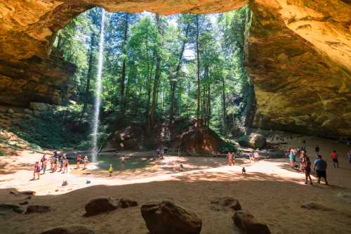 A scenic cave with a waterfall, surrounded by lush trees and people enjoying the sandy area below.