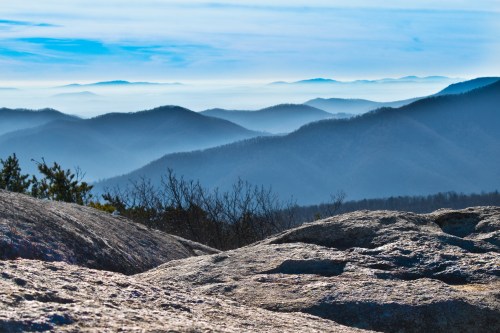 A panoramic view of layered blue mountains under a clear sky, with rocky foreground and soft mist in the valleys.