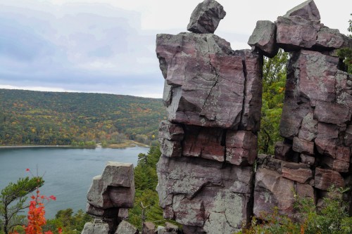 Rock formations overlook a serene lake surrounded by autumn foliage and rolling hills under a cloudy sky.
