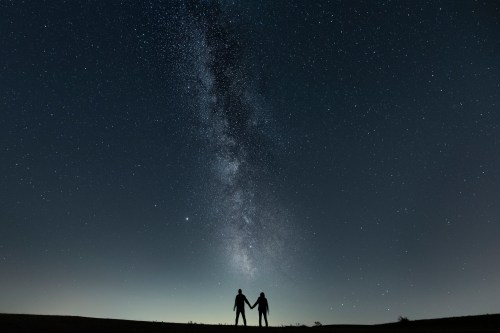 Silhouetted couple holding hands under a starry sky with the Milky Way stretching above them.