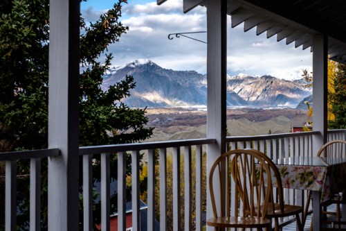 A scenic view from a porch, featuring mountains and valleys under a partly cloudy sky. Two wooden chairs are visible.