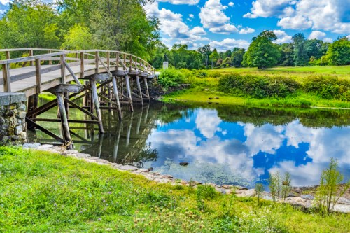 A wooden bridge over a calm river reflects fluffy clouds and greenery in a serene landscape.