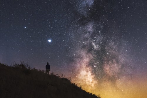 A silhouette of a person stands on a hill, gazing at a starry sky filled with the Milky Way and bright celestial bodies.