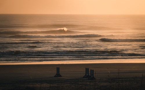 A serene beach scene at sunset, with gentle waves and a lone surfer riding a wave in the distance.