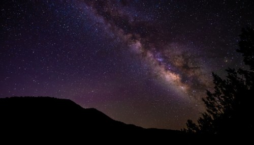 A stunning view of the Milky Way galaxy over a dark silhouette of mountains and trees against a starry night sky.