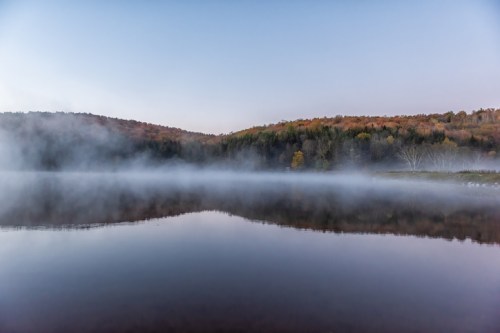 A serene lake at dawn, surrounded by mist and autumn-colored trees reflecting on the calm water.