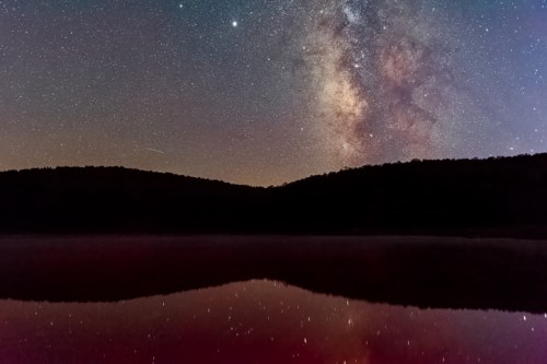 A serene night sky filled with stars and the Milky Way, reflecting on a calm lake surrounded by dark hills.