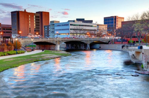 A river flows through a cityscape at sunset, with buildings and a bridge in the background.