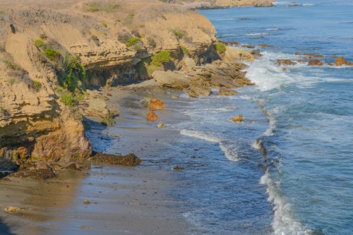 Coastal landscape with rocky cliffs, sandy beach, and gentle waves lapping at the shore under a clear blue sky.
