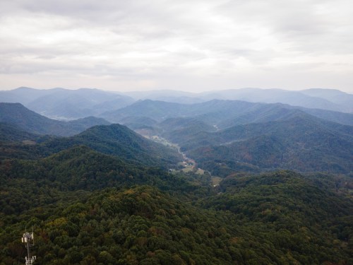 Aerial view of rolling green mountains under a cloudy sky, with valleys and a small village visible in the distance.