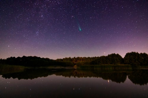 A serene night sky filled with stars and a comet, reflecting on a calm lake surrounded by trees.