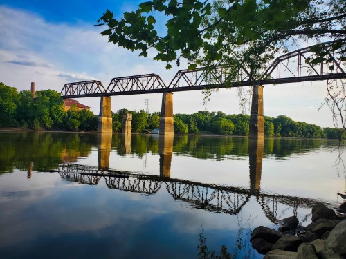 A bridge spans a calm river, reflecting trees and blue sky, with a peaceful landscape in the background.